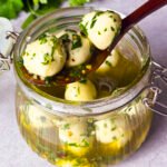 GLASS JAR, FILLED WITH MOZZARELLA BALLS AND OLIVE OILE MARINADE. WOODEN SPOON IS RESTING ON THE RIM WITH 3 MOZZARELLA BALLS.