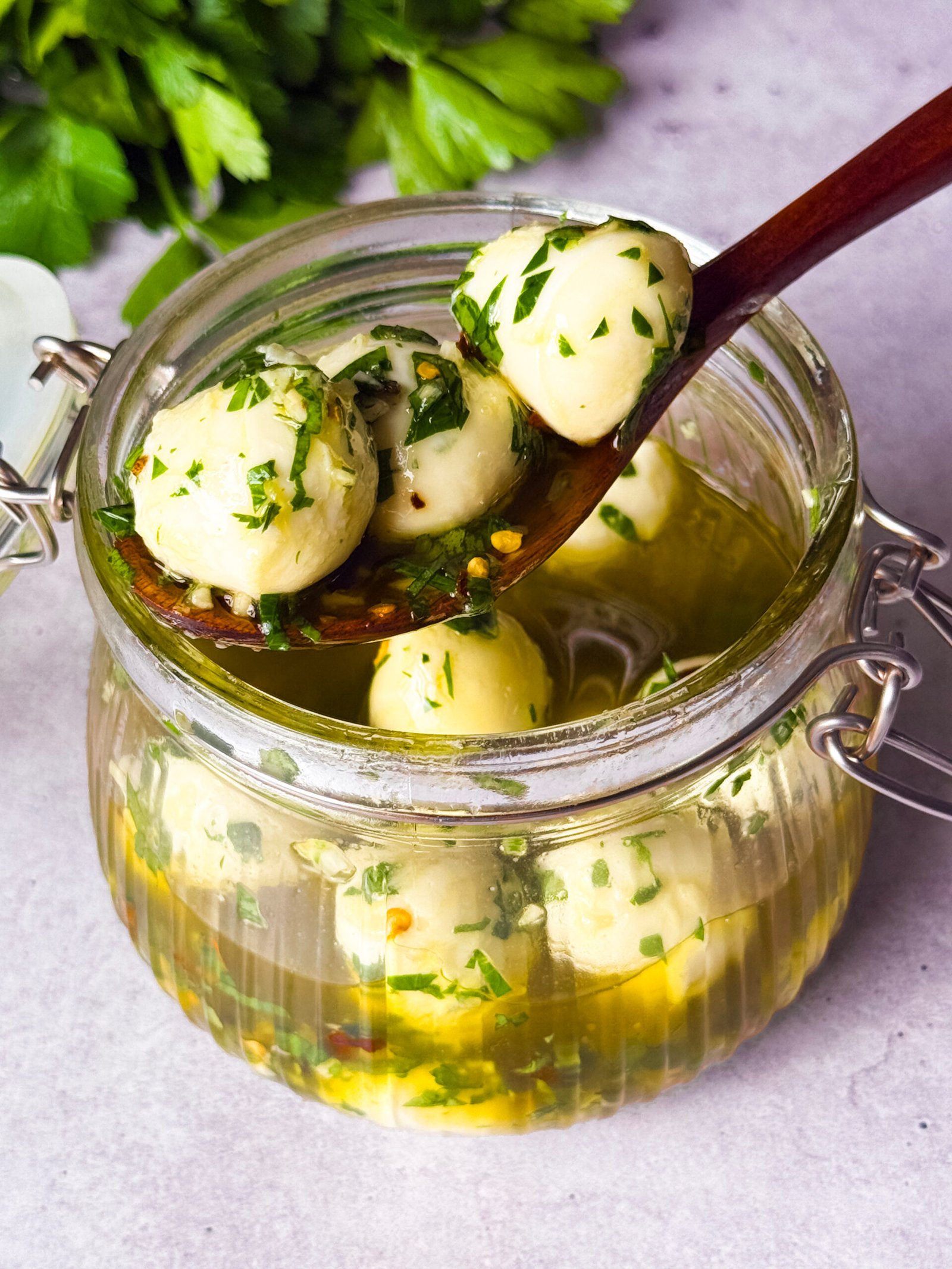 GLASS JAR, FILLED WITH MOZZARELLA BALLS AND OLIVE OILE MARINADE. WOODEN SPOON IS RESTING ON THE RIM WITH 3 MOZZARELLA BALLS.