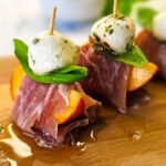 Slices of peach, wrapped with prosciutto wit basil and mozzarella balls on a wooden board.