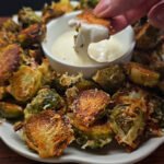 a plate with roasted Brussels sprouts. Hand is holding one of the sprouts, dipped in the sauce.