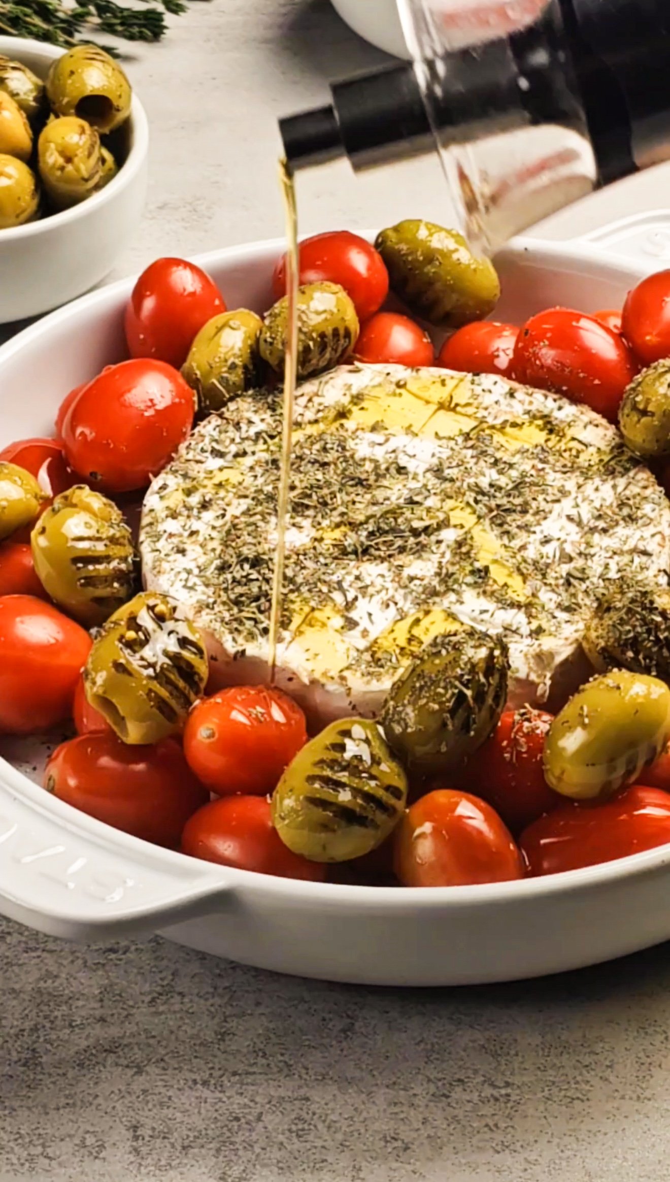 Brie cheese with olives and tomatoes in a baking dish. Olive oil is being drizzled on top.