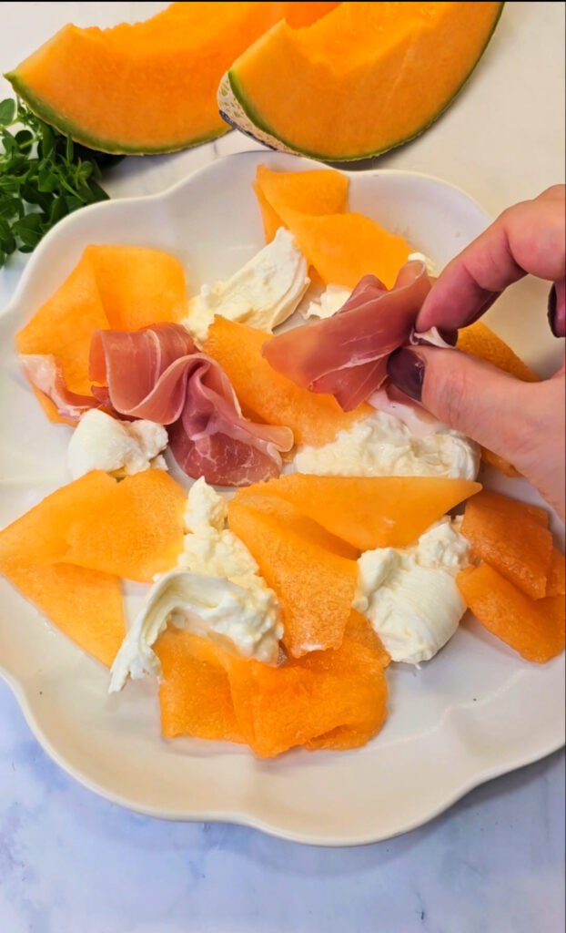 Burrata, melon and prosciutto on a plate, hand is placing prosciutto on the same plate