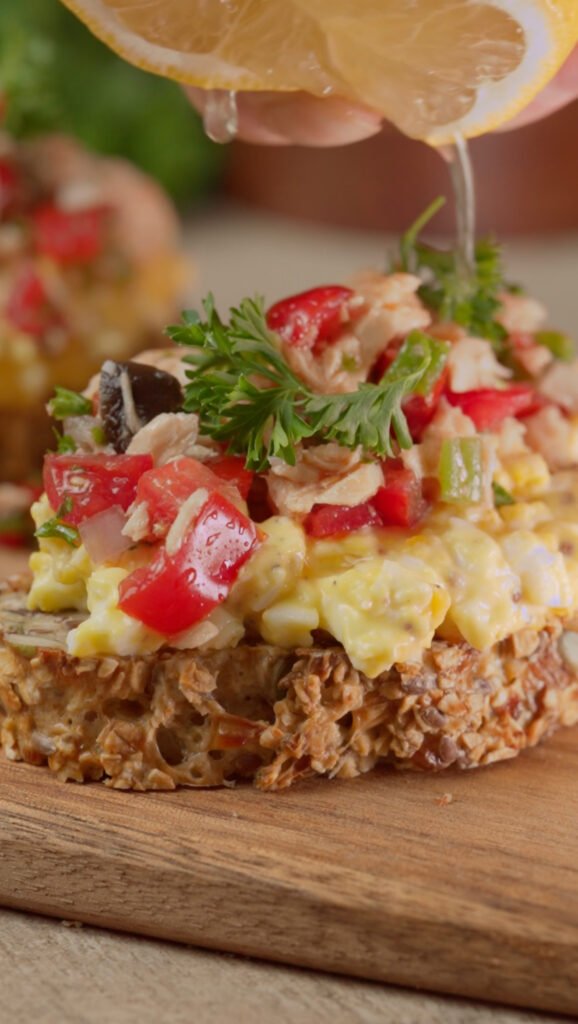Layered Tuna and Egg Salad Toast on a wooden board. Lemon juice is being squeezed on top