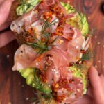 Two hands are holding the toast with avocado and prosciutto
