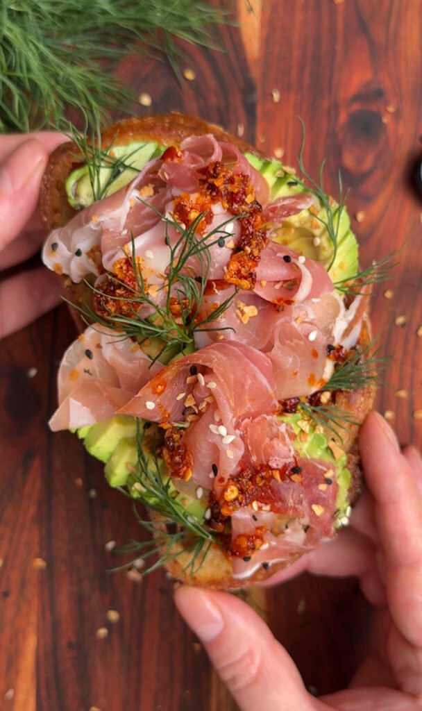Two hands are holding the toast with avocado and prosciutto