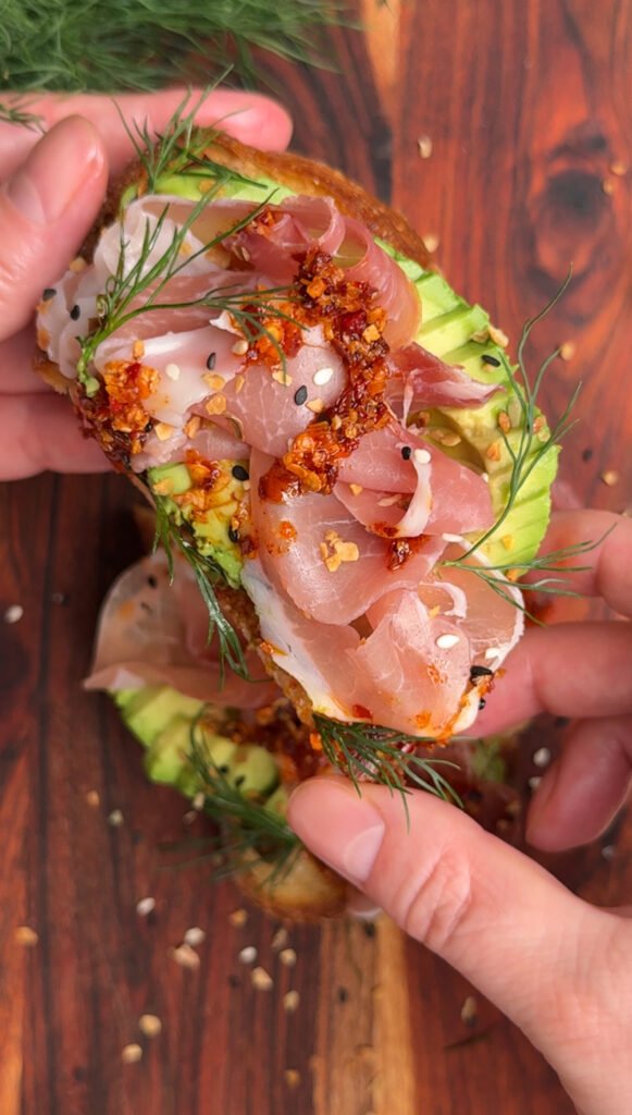hands are holding the sliced piece of toast with avocado and prosciutto