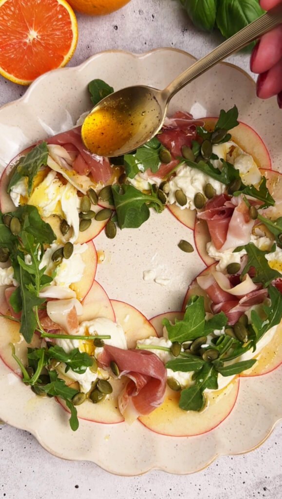 A golden spoon drizzling orange honey dressing over apple slices, burrata, prosciutto, arugula, and pumpkin seeds on a platter.