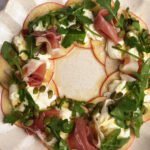 Close-up of a beautiful apple burrata and prosciutto salad wreath, topped with fresh arugula and pumpkin seeds, drizzled with orange honey dressing.