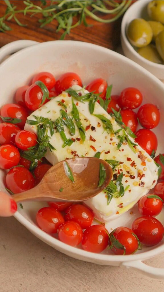 Tomatoes and cream cheese in a baking dish. Wooden spoon is spreading herbs and oil on top