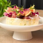 Goat Cheese & Fig Stack appetizer layered with prosciutto, figs, and hot honey on a white pedestal stand