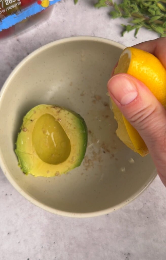 avocado half in a bowl, hand is squeezing the lemon