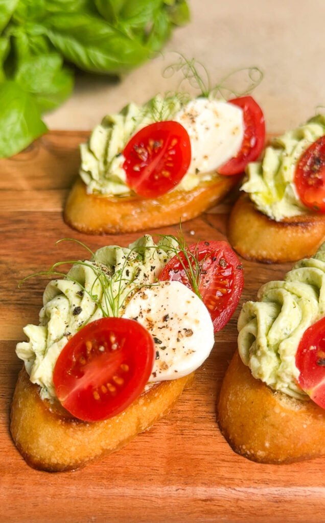 Caprese-inspired crostini topped with pesto cream cheese, cherry tomatoes, and fresh mozzarella on a wooden board with fresh basil in the background.