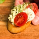 Close-up of a hand holding a toasted crostini topped with piped pesto cream cheese, halved cherry tomato, mozzarella slice, and microgreens.