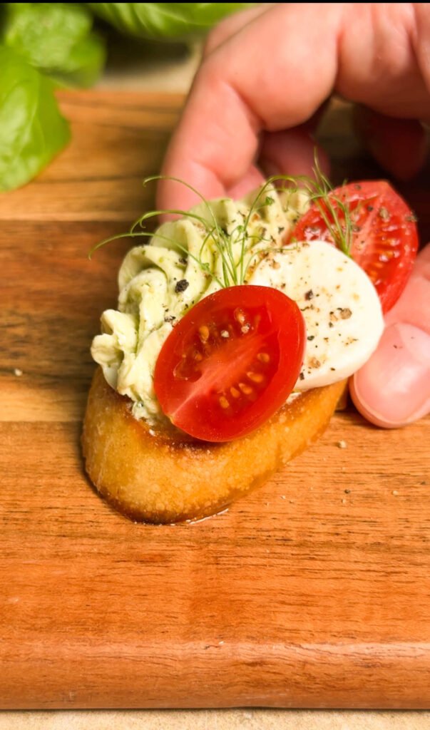 Close-up of a hand holding a toasted crostini topped with piped pesto cream cheese, halved cherry tomato, mozzarella slice, and microgreens.