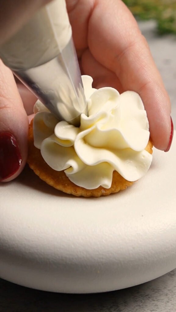Close-up of a hand piping creamy whipped goat cheese onto a buttery Ritz cracker using a pastry bag with a star tip