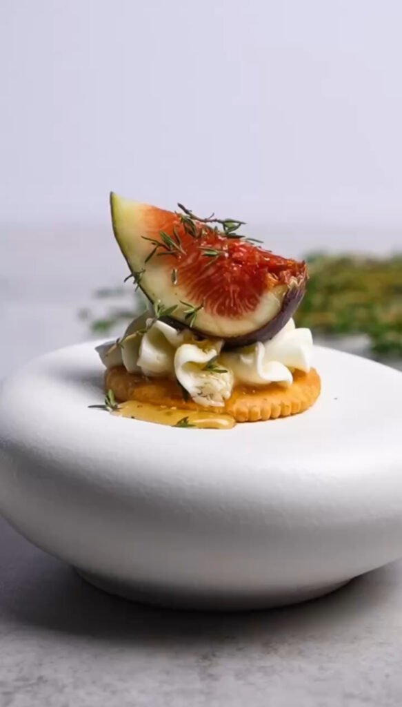 Elegant honey whipped goat cheese and fresh fig bite on a Ritz cracker, garnished with thyme and honey drizzle, styled on a white pedestal plate.