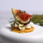 Elegant honey whipped goat cheese and fresh fig bite on a Ritz cracker, garnished with thyme and honey drizzle, styled on a white pedestal plate.