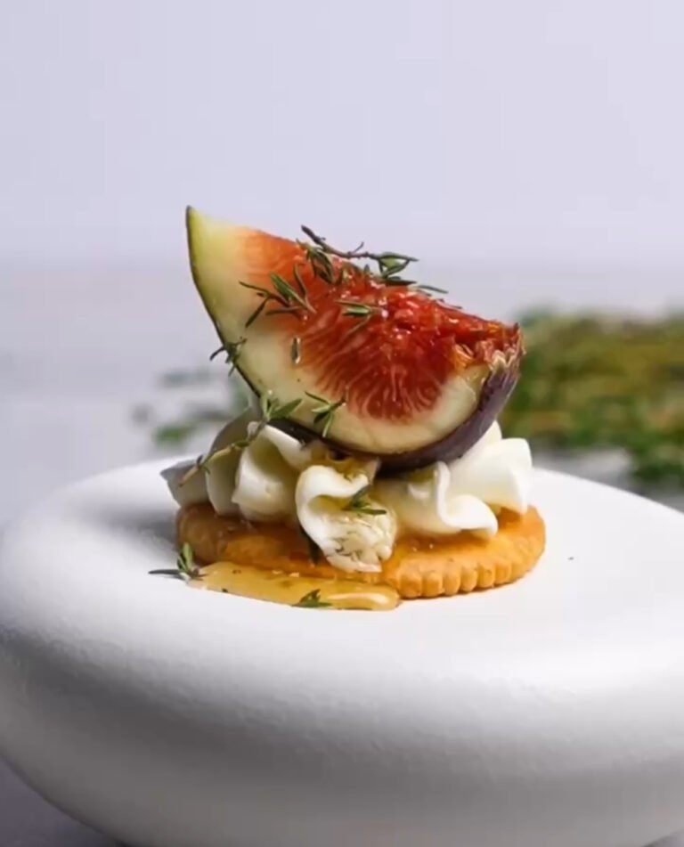 Elegant honey whipped goat cheese and fresh fig bite on a Ritz cracker, garnished with thyme and honey drizzle, styled on a white pedestal plate.