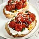 Close-up of golden toasted bread topped with creamy labneh, honey roasted cherry tomatoes, freshly grated parmesan, and sprinkled with herbs and nigella seeds on a white plate
