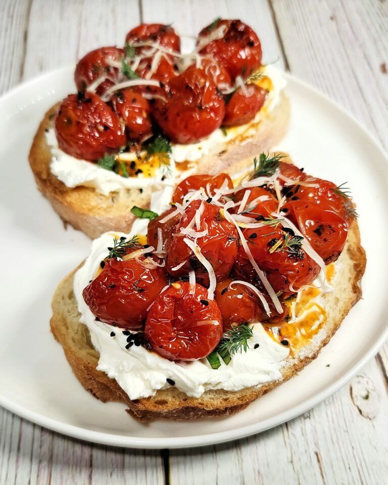 Close-up of golden toasted bread topped with creamy labneh, honey roasted cherry tomatoes, freshly grated parmesan, and sprinkled with herbs and nigella seeds on a white plate