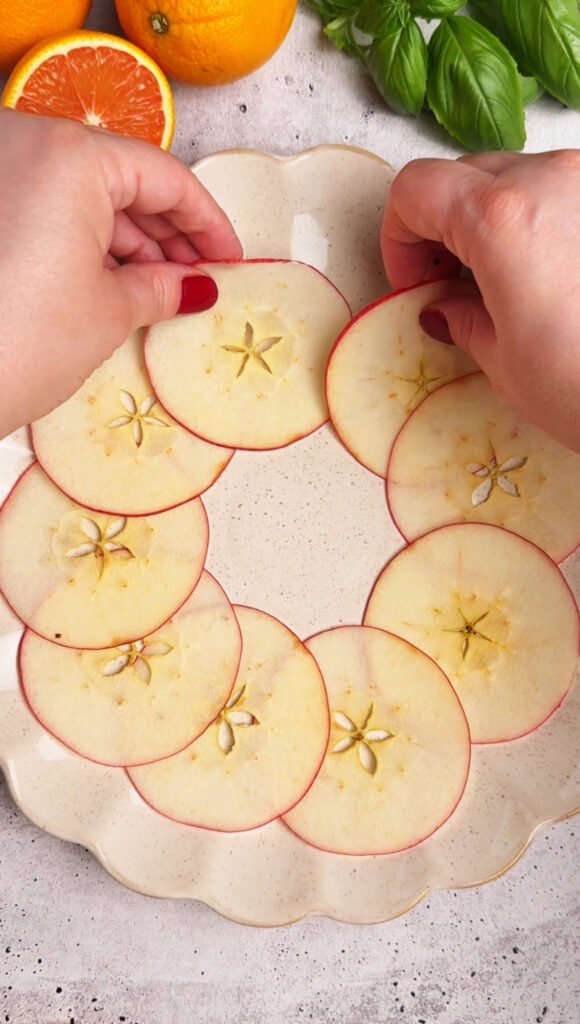 Hands arranging thin apple slices in a circular pattern on a cream-colored platter to create the base for an apple burrata and prosciutto salad wreath.