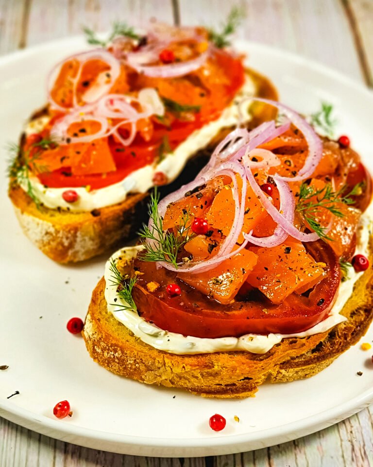 Two slices of golden toasted sourdough topped with cream cheese, fresh tomato slices, and vibrant salmon tartare, garnished with thin shallot rings, dill, and pink peppercorns on a white plate.