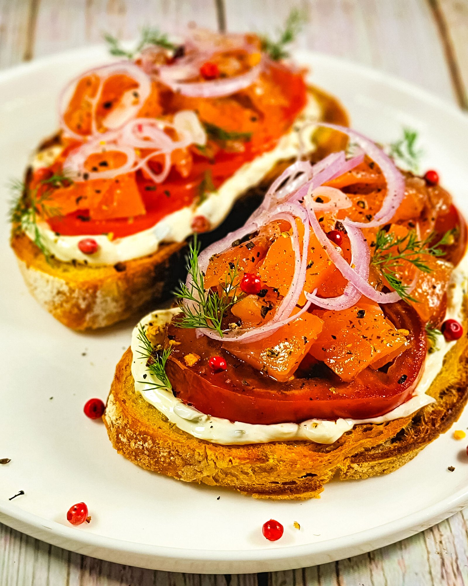 Two slices of golden toasted sourdough topped with cream cheese, fresh tomato slices, and vibrant salmon tartare, garnished with thin shallot rings, dill, and pink peppercorns on a white plate.