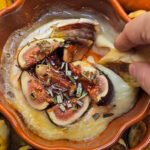Baked Brie topped with figs, plum jelly, and rosemary in an orange pumpkin-shaped dish, surrounded by toasted bread slices, with a hand dipping into the gooey cheese