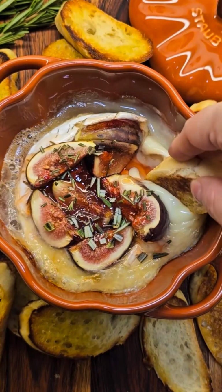 Baked Brie topped with figs, plum jelly, and rosemary in an orange pumpkin-shaped dish, surrounded by toasted bread slices, with a hand dipping into the gooey cheese