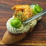 A close-up of a Cajun Shrimp Crostini on a wooden board. The toasted baguette slice is topped with piped shrimp mousse, a golden Cajun-spiced shrimp, a delicate cucumber ribbon, and a sprig of fresh dill