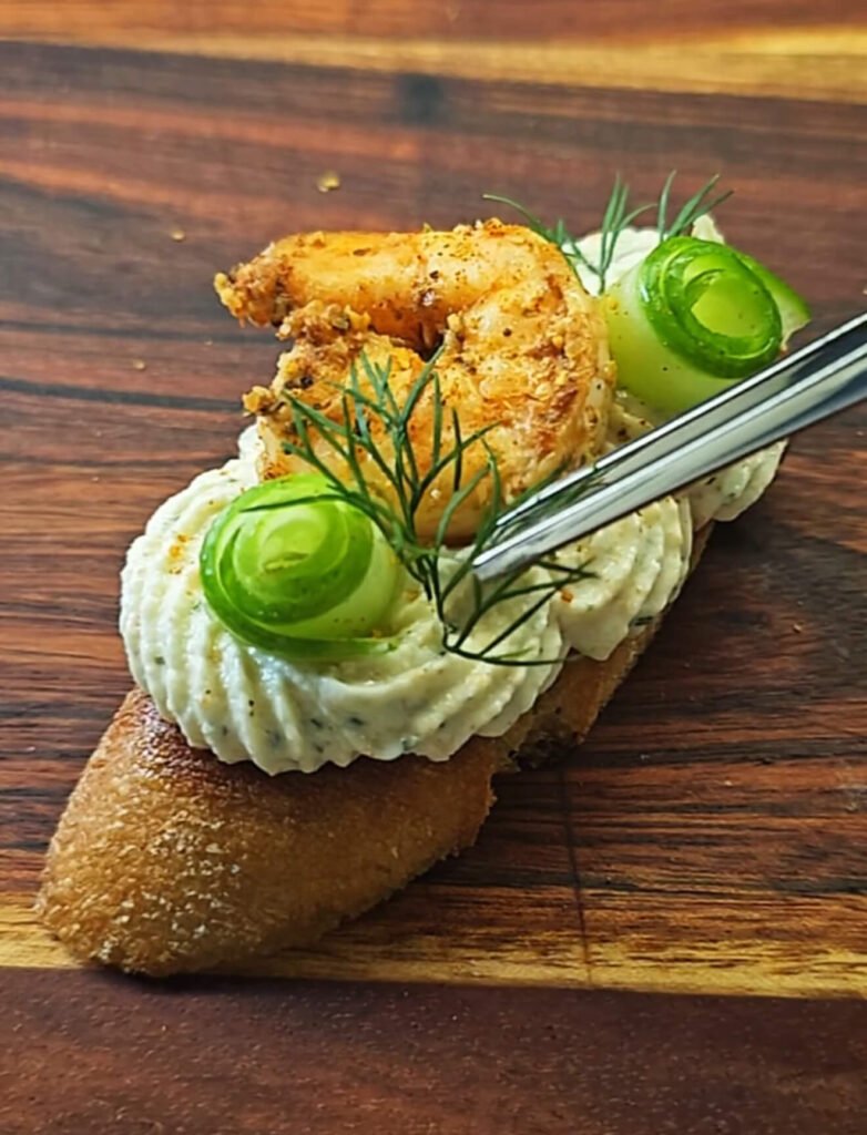 A close-up of a Cajun Shrimp Crostini on a wooden board. The toasted baguette slice is topped with piped shrimp mousse, a golden Cajun-spiced shrimp, a delicate cucumber ribbon, and a sprig of fresh dill