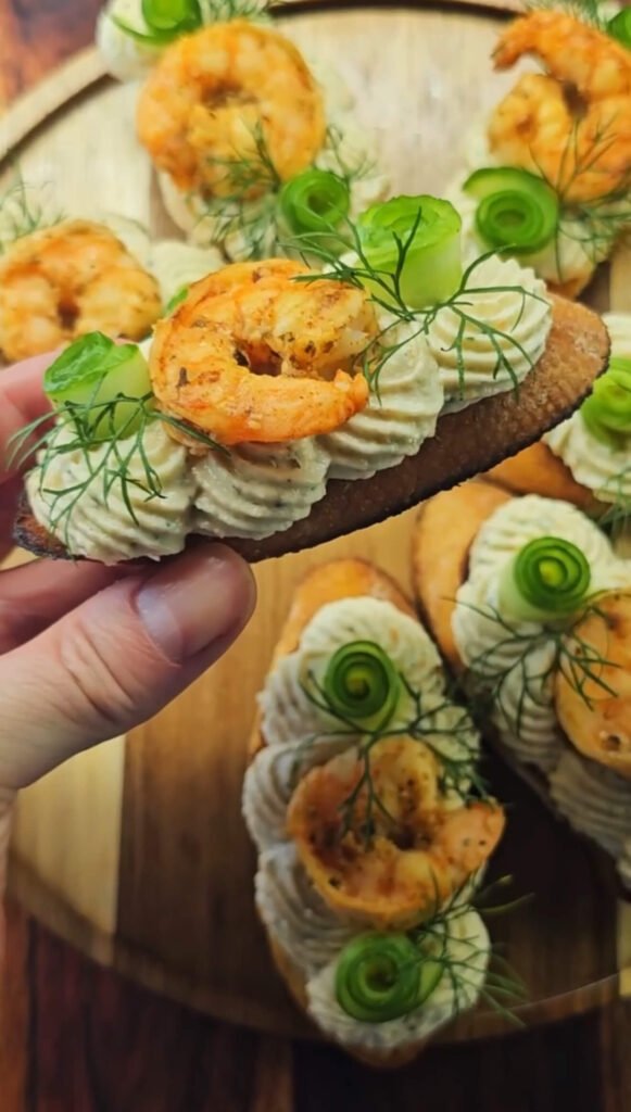 A hand holding one Cajun Shrimp Crostini topped with creamy shrimp mousse, a seasoned shrimp, cucumber ribbon, and fresh dill. Several more crostinis are arranged on a round wooden platter in the background.
