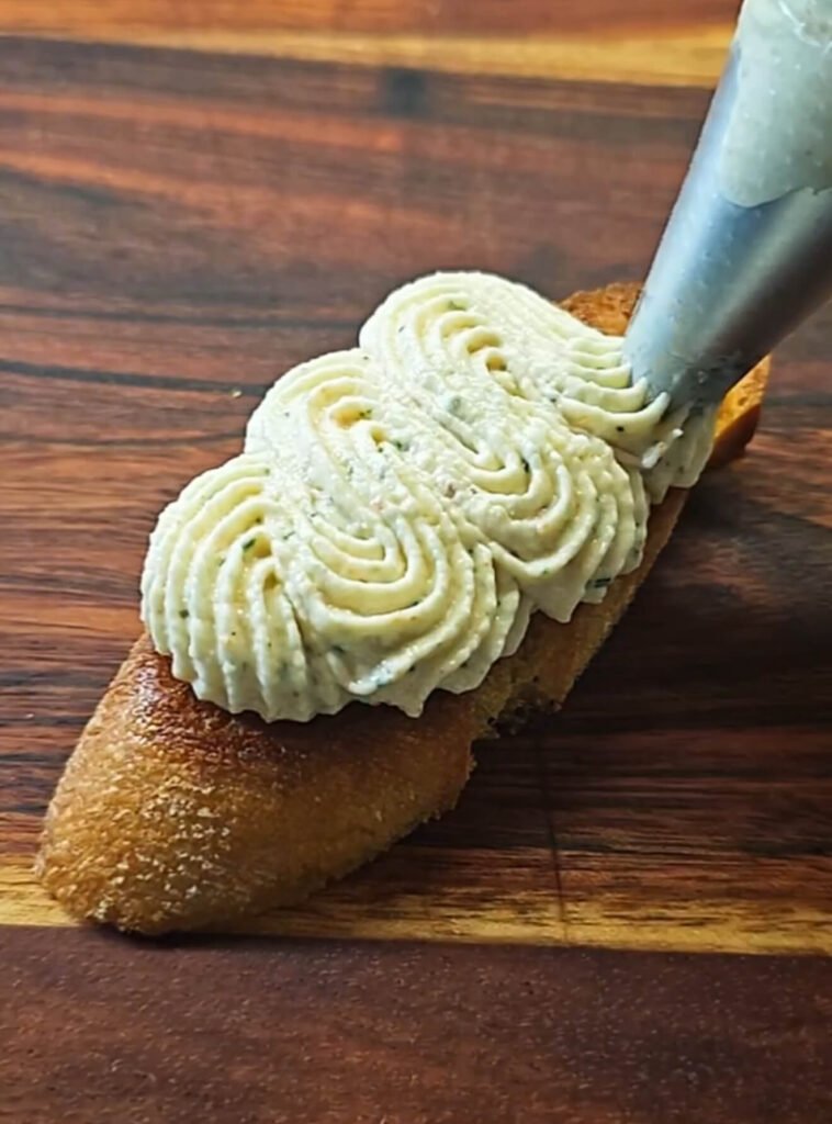 A toasted baguette slice being topped with piped shrimp mousse from a pastry bag, showing a detailed close-up of the smooth, creamy mousse swirls on the crisp bread.