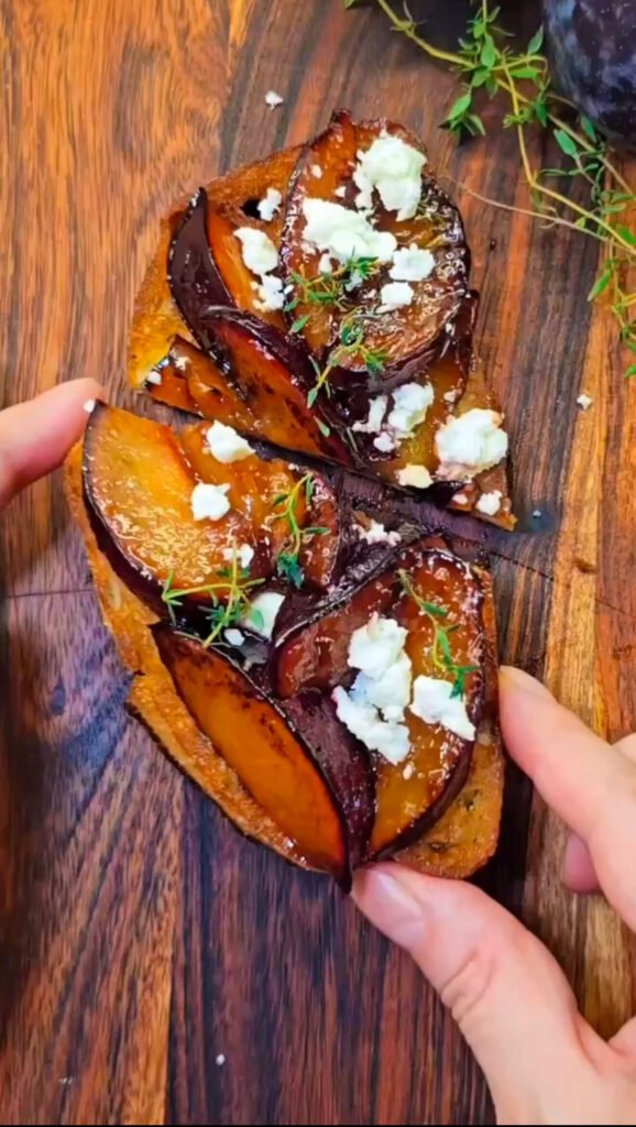 Close-up of a slice of toasted bread topped with caramelized plum wedges, crumbled feta cheese, and fresh thyme, served on a wooden board.