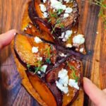 Close-up of a slice of toasted bread topped with caramelized plum wedges, crumbled feta cheese, and fresh thyme, served on a wooden board.