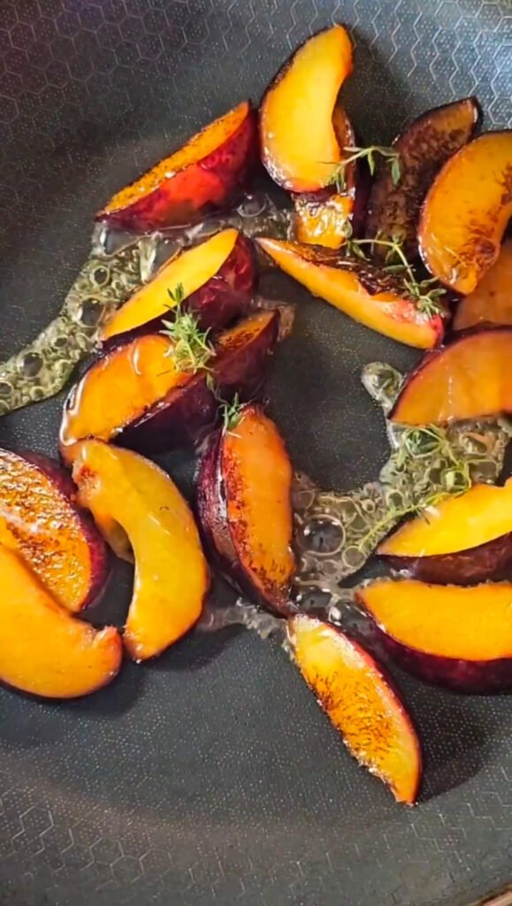 Sliced plums caramelizing in a pan with honey and fresh thyme sprigs, bubbling in syrup.