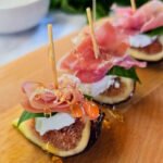 Close-up of fig and cheese bites on a wooden board, made with fresh fig quarters topped with creamy goat cheese, basil, folded prosciutto, and drizzled with golden honey.
