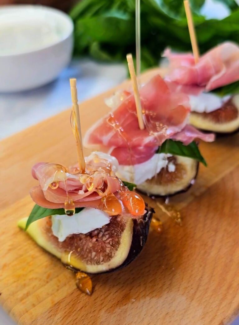 Close-up of fig and cheese bites on a wooden board, made with fresh fig quarters topped with creamy goat cheese, basil, folded prosciutto, and drizzled with golden honey.