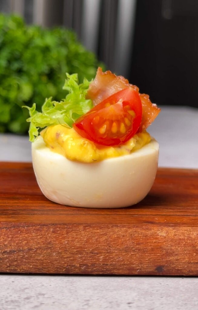 Single BLT deviled egg on a wooden serving board, filled with creamy yolk mixture and topped with a small green lettuce leaf, a quartered cherry tomato, and a crisp bacon piece, with a soft-focus kitchen background.