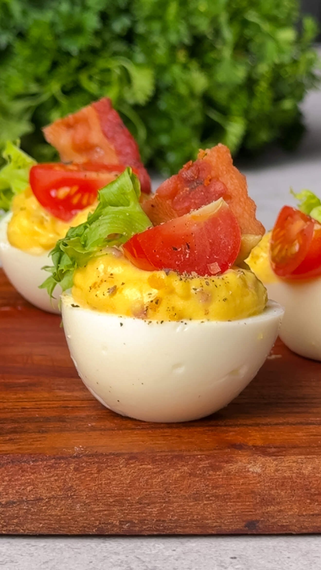 Close-up of a BLT deviled egg on a wooden serving board, topped with a quartered cherry tomato, a small piece of crispy bacon, and a ruffled piece of green lettuce, with a blurred background of fresh parsley.