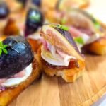 Close-up of a brie crostini topped with fig jam, melted brie cheese, a slice of prosciutto, and a fresh fig quarter, drizzled with honey and garnished with a small thyme sprig, served on a wooden board.
