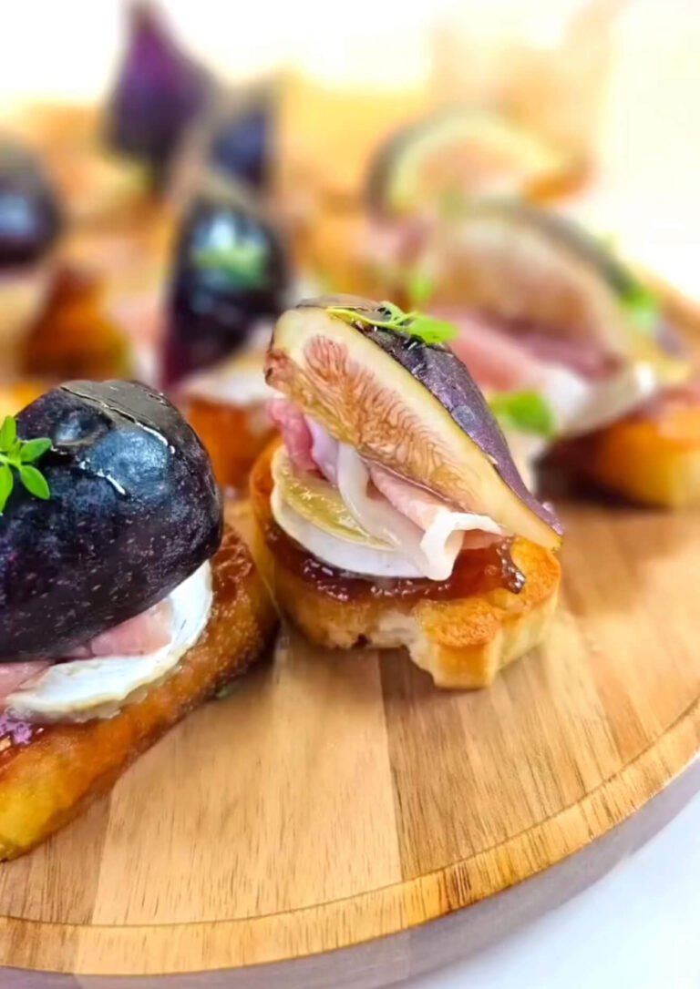 Close-up of a brie crostini topped with fig jam, melted brie cheese, a slice of prosciutto, and a fresh fig quarter, drizzled with honey and garnished with a small thyme sprig, served on a wooden board.