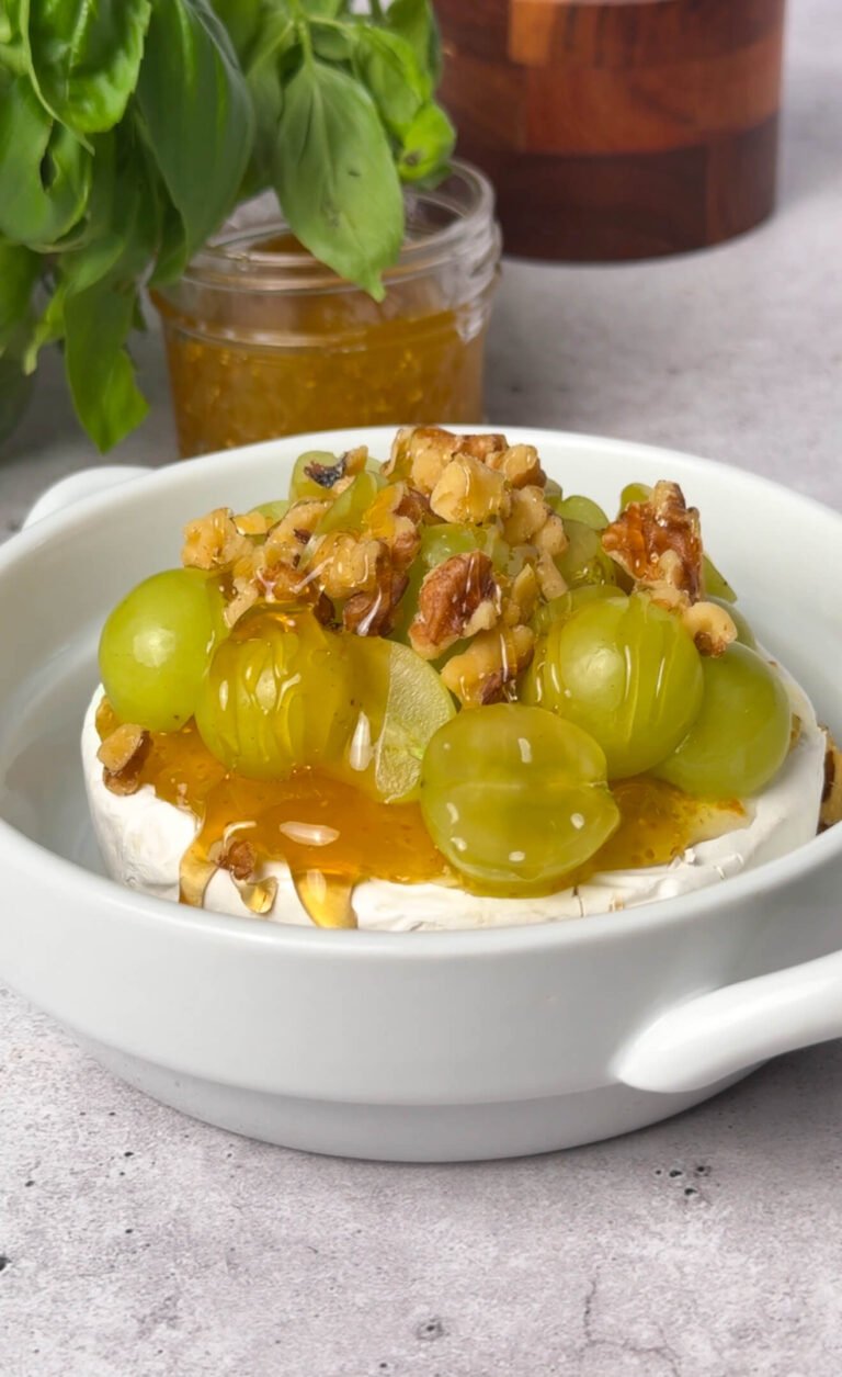 Overhead shot of Brie cheese topped with halved grapes, chopped walnuts, and honey drizzle, ready to be baked in a white dish