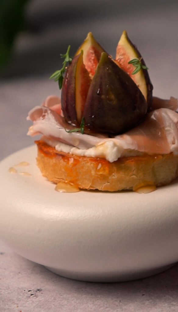 Caramelized fig and prosciutto crostini on a toasted baguette slice, topped with whipped ricotta, fresh thyme, chopped walnuts, and a drizzle of honey, served as an elegant appetizer.