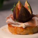 Caramelized fig and prosciutto crostini on a toasted baguette slice, topped with whipped ricotta, fresh thyme, chopped walnuts, and a drizzle of honey, served as an elegant appetizer.