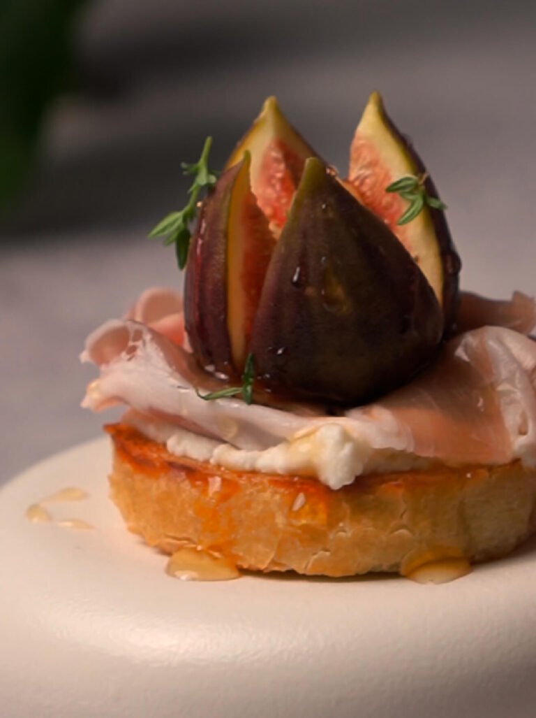 Caramelized fig and prosciutto crostini on a toasted baguette slice, topped with whipped ricotta, fresh thyme, chopped walnuts, and a drizzle of honey, served as an elegant appetizer.