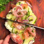 Cucumber ribbon and prosciutto toast on sourdough being sliced, topped with lemon-ricotta, capers, and thinly sliced shallots, with fresh parsley and lemon in the background.