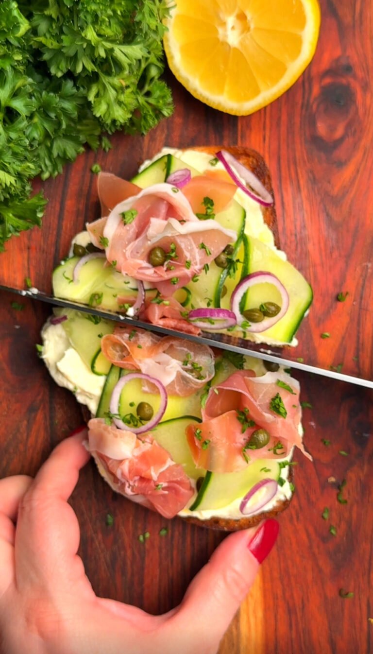 Cucumber ribbon and prosciutto toast on sourdough being sliced, topped with lemon-ricotta, capers, and thinly sliced shallots, with fresh parsley and lemon in the background.
