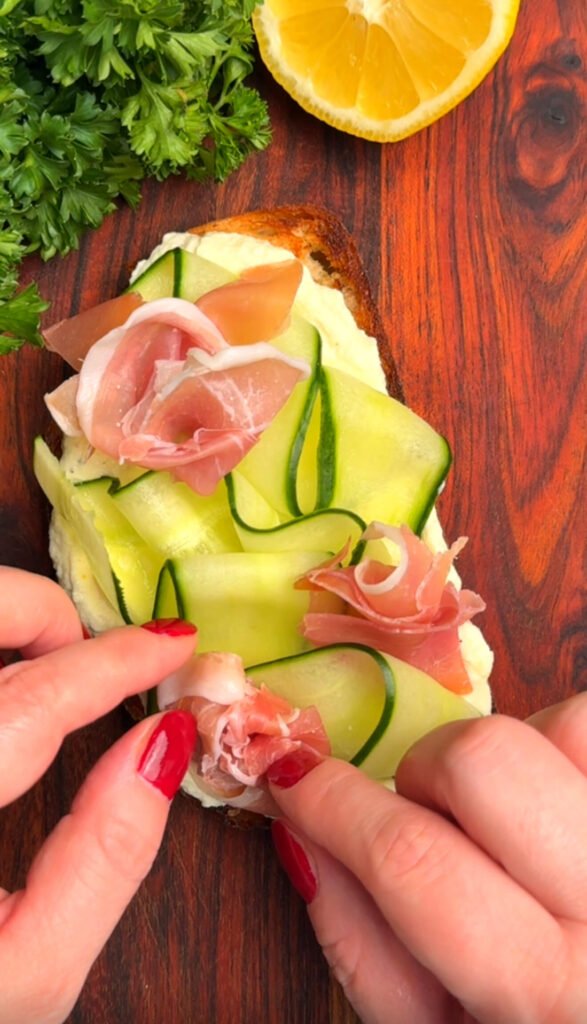 Hands arranging prosciutto roses on top of cucumber ribbon and lemon-ricotta toast, with fresh parsley and lemon in the corner of the frame.