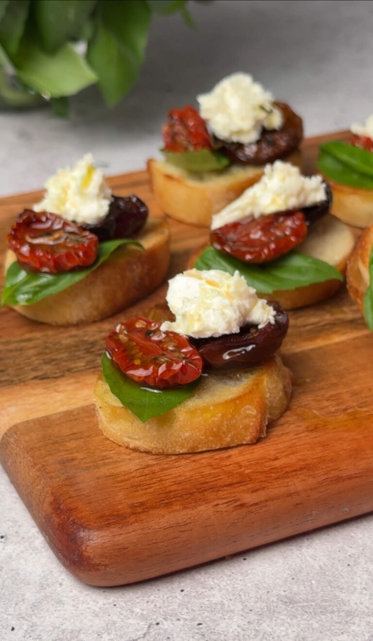 oasted baguette crostini topped with fresh basil, homemade sun-dried tomato with plum, and a chunk of creamy feta cheese, drizzled with olive oil, served on a wooden board.